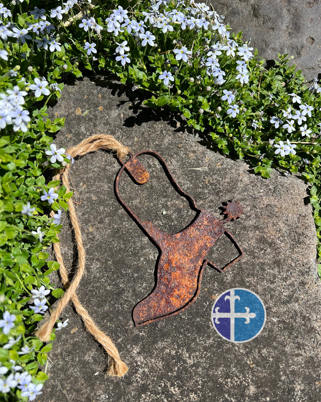 A rusted steel cowboy boot ornament with a spur design, laying on a stone surface surrounded by small white flowers and greenery. The ornament features a natural, weathered rust patina, with a coarse rope loop attached for hanging. A circular logo with a cross and fleur-de-lis design is visible near the bottom right corner of the image.