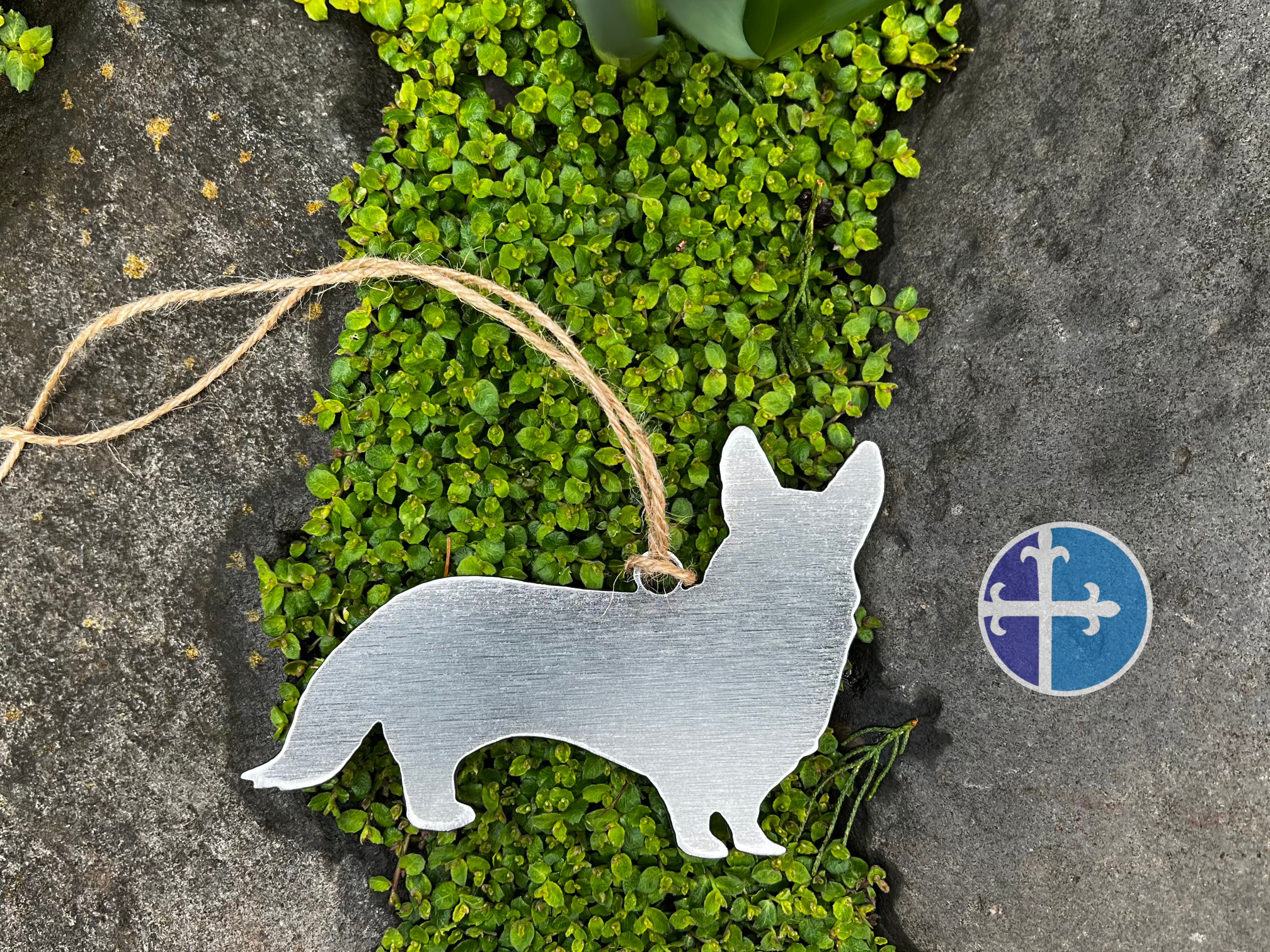 Handmade steel Corgi ornament, rustic twine loop, laid on lush greenery and stone.