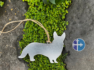 Handmade steel Corgi ornament, rustic twine loop, laid on lush greenery and stone.