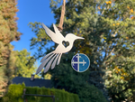 Load image into Gallery viewer, Metal art hummingbird ornament, suspended in a sunny outdoor scene, showcasing the heart cutout.
