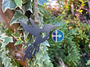 Hummingbird-shaped metal ornament with a rustic finish, displayed on ivy-covered wood.
