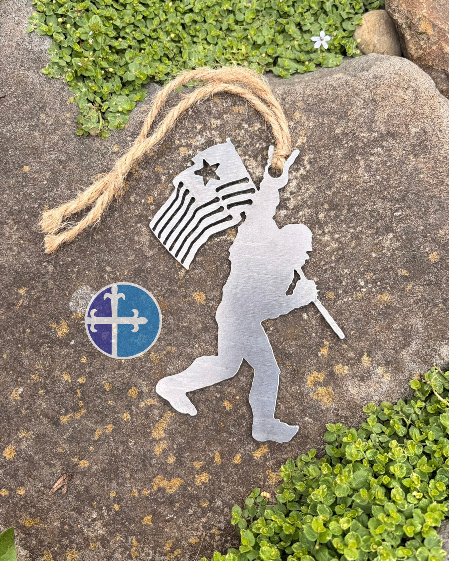 Steel Bigfoot ornament carrying an American flag with a rock-on hand gesture resting on a stone surrounded by green ground cover.