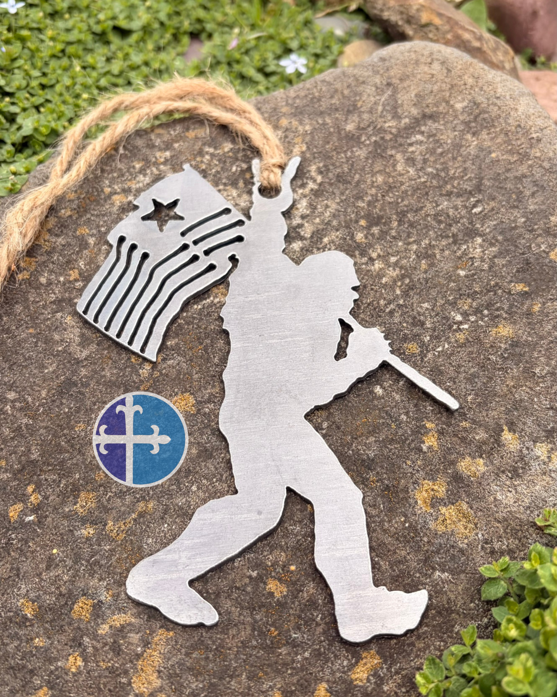 Patriotic Bigfoot metal ornament displayed on a large stone with green plants and natural outdoor background.