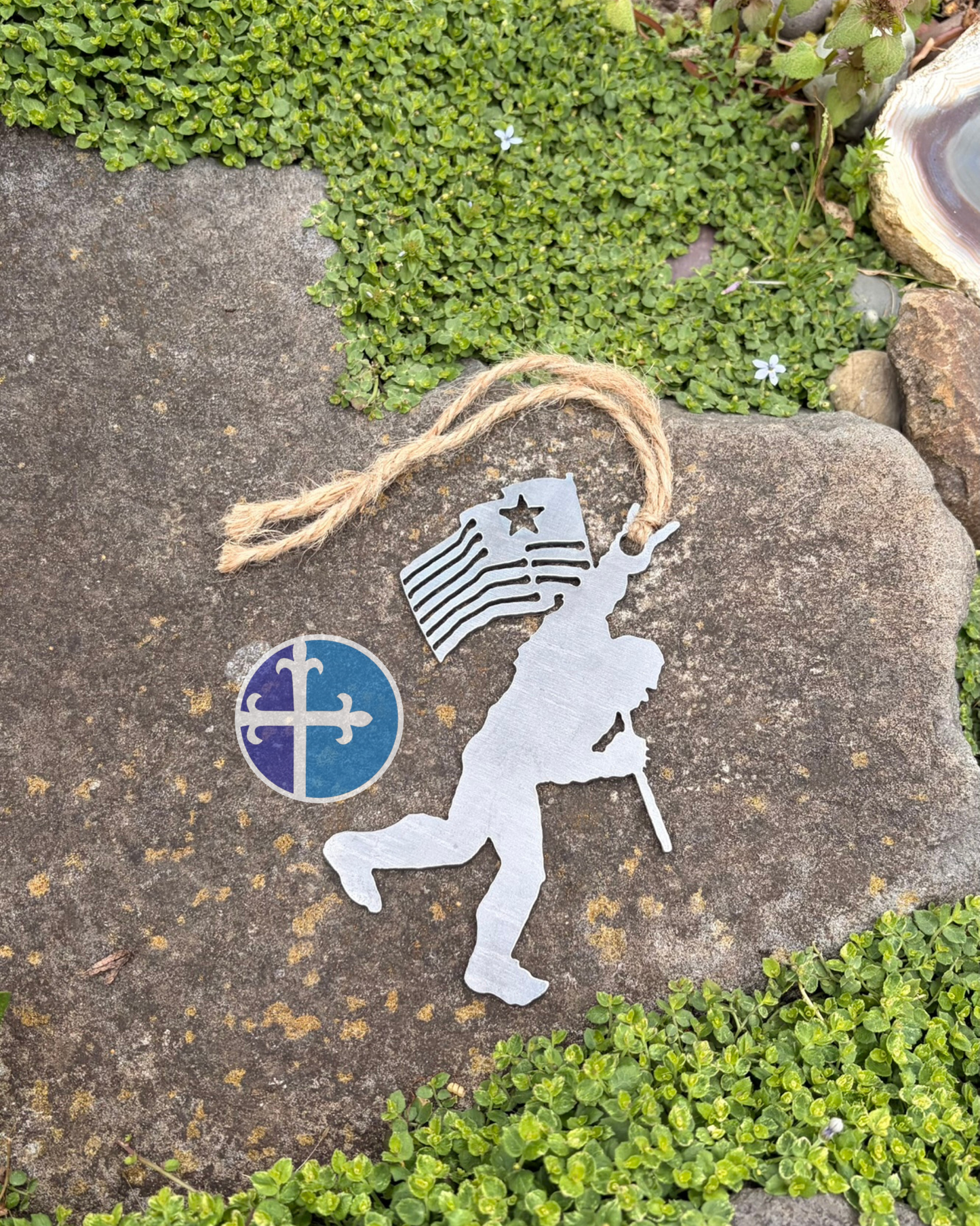 Bigfoot steel Christmas ornament with American flag and raised rock-on hand sign photographed on a flat stone surface.