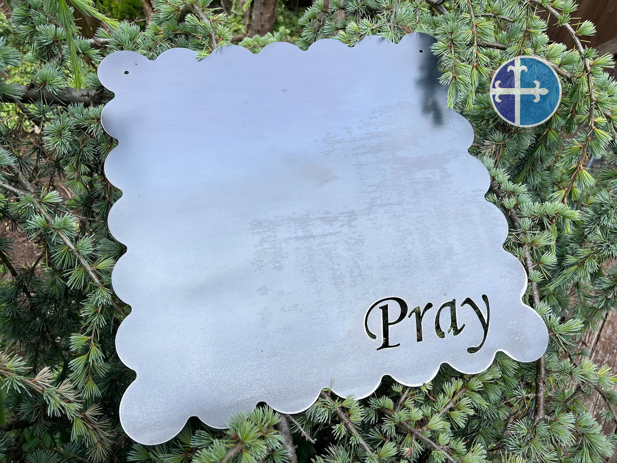 Raw steel scalloped magnet board with Pray cutout displayed against green evergreen branches.