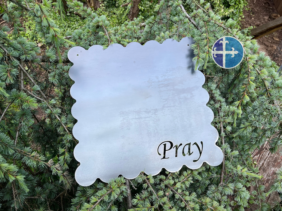 Angled close-up of a raw steel scalloped magnet board with Pray cutout and evergreen background.