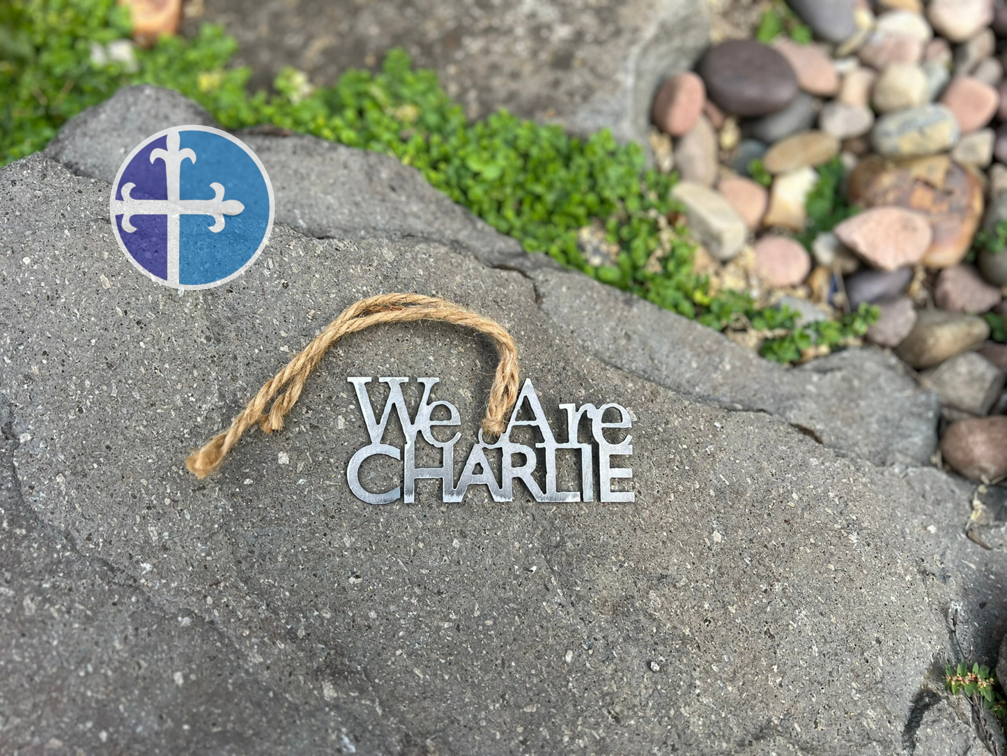 Steel ornament reading “We Are Charlie” on a stone surface with rustic jute twine.