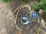 Load image into Gallery viewer, Steaming steel coffee cup ornament with rustic twine, lying on a textured stone surface surrounded by greenery.
