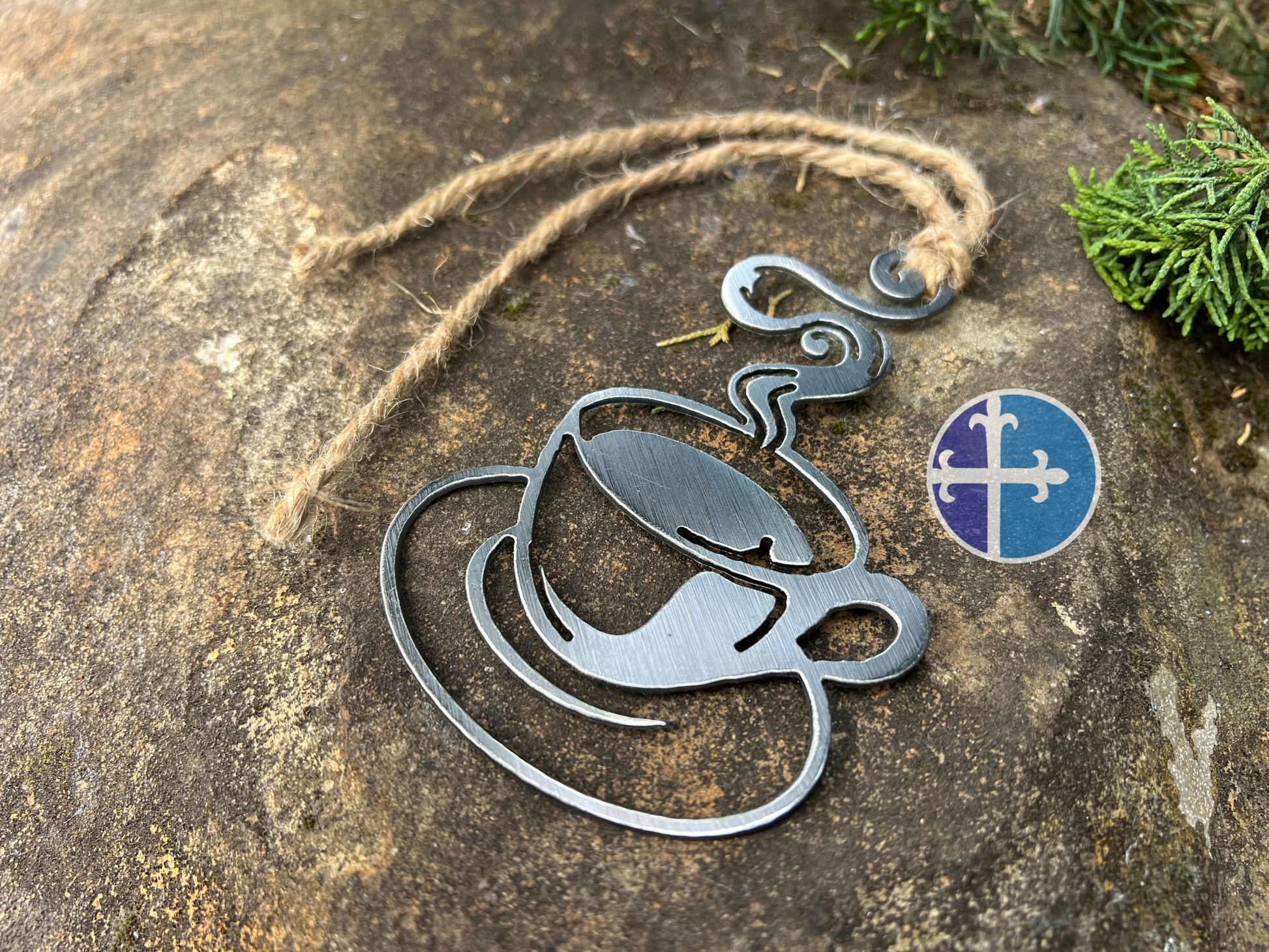 Close-up view of a steaming steel coffee cup ornament with a rustic twine hanging loop, placed on a stone surface with green plants in the background.
