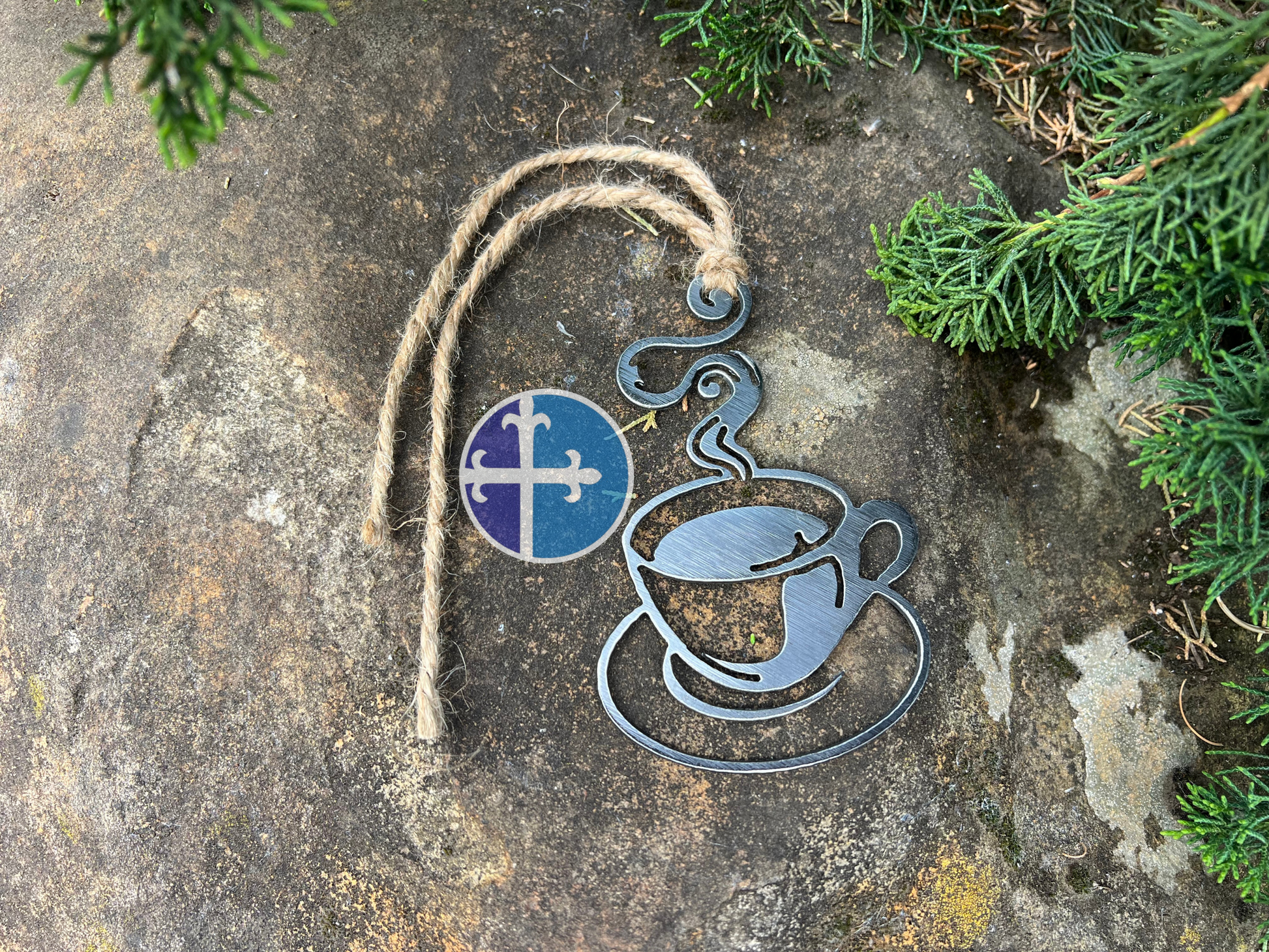 Steaming steel coffee cup ornament hanging by a rustic twine on a stone surface, featuring industrial-style details and surrounded by natural greenery.