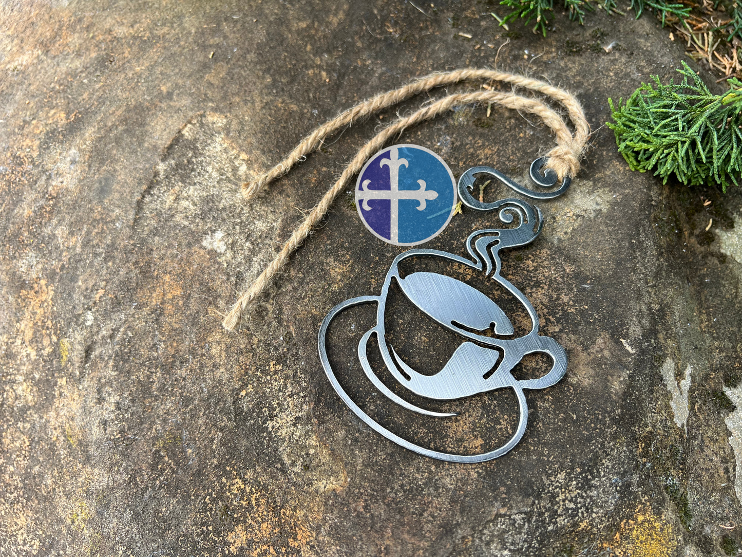 Handmade steaming steel coffee cup ornament with rustic twine, displayed on a rock with green foliage in the background.