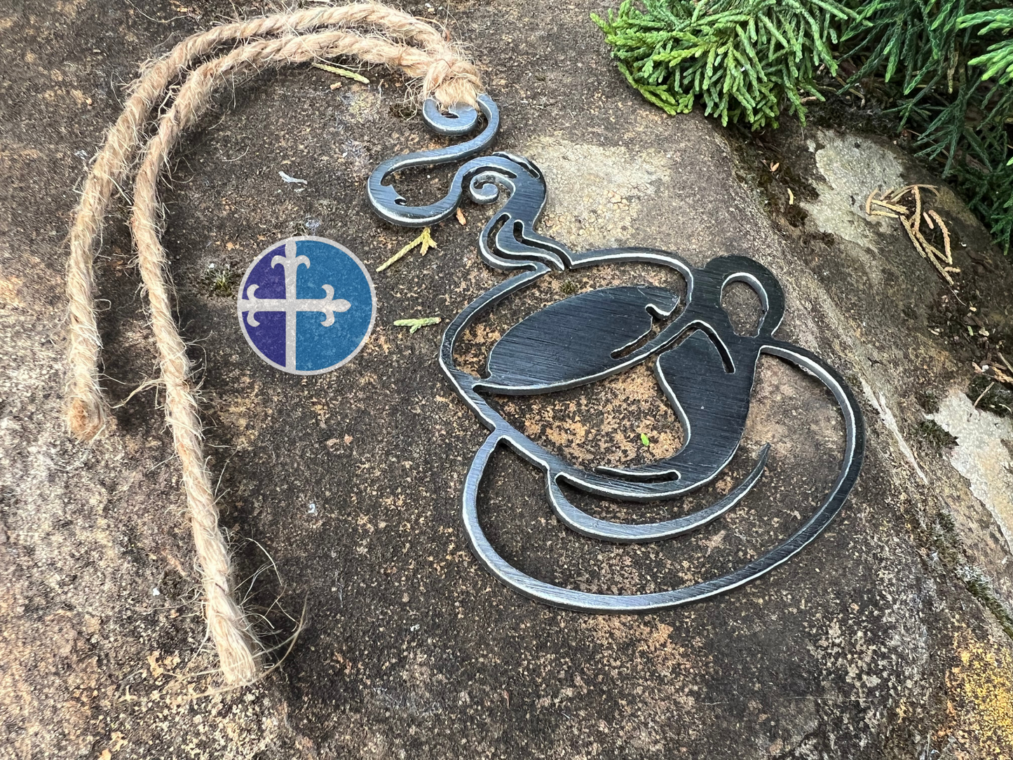 Steaming steel coffee cup ornament hanging by rustic twine on a textured stone surface, showcasing industrial design elements with green plants nearby.
