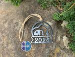 Load image into Gallery viewer, A metal 2024 camper ornament with a mountain and tree design, hanging by a rustic twine on a natural background.

