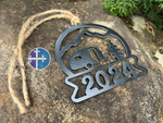 Load image into Gallery viewer, Side view of the 2024 camper Christmas ornament, made from steel, featuring a mountain and RV design, laid on a rustic surface.
