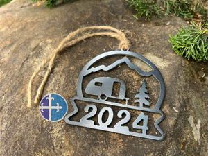 Close-up of the 2024 camper ornament with a detailed cutout of an RV and tree against a mountain backdrop, hung by twine.