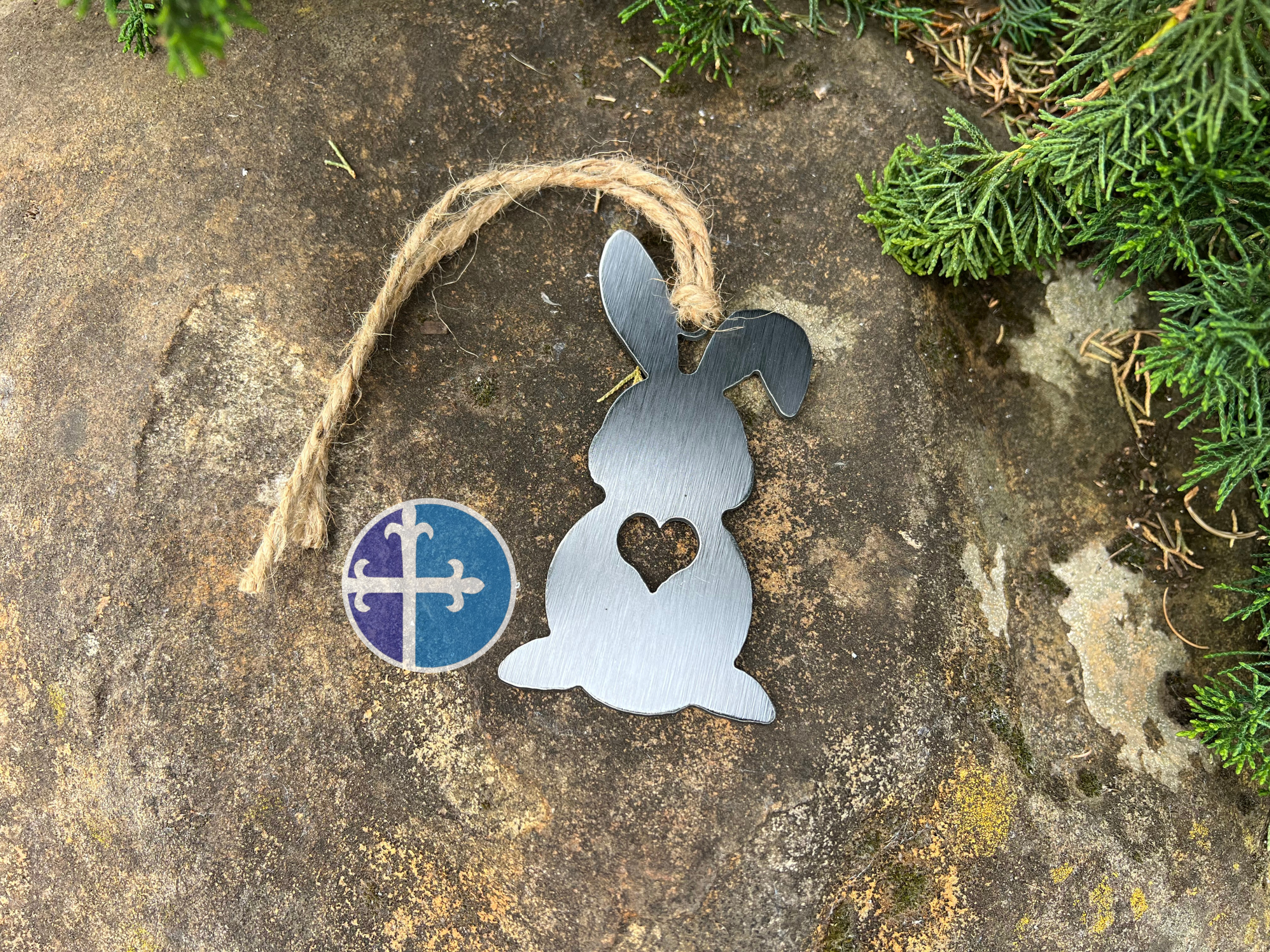 Overhead view of a steel bunny silhouette ornament with a heart cutout, displayed on a stone surface with greenery in the background.
