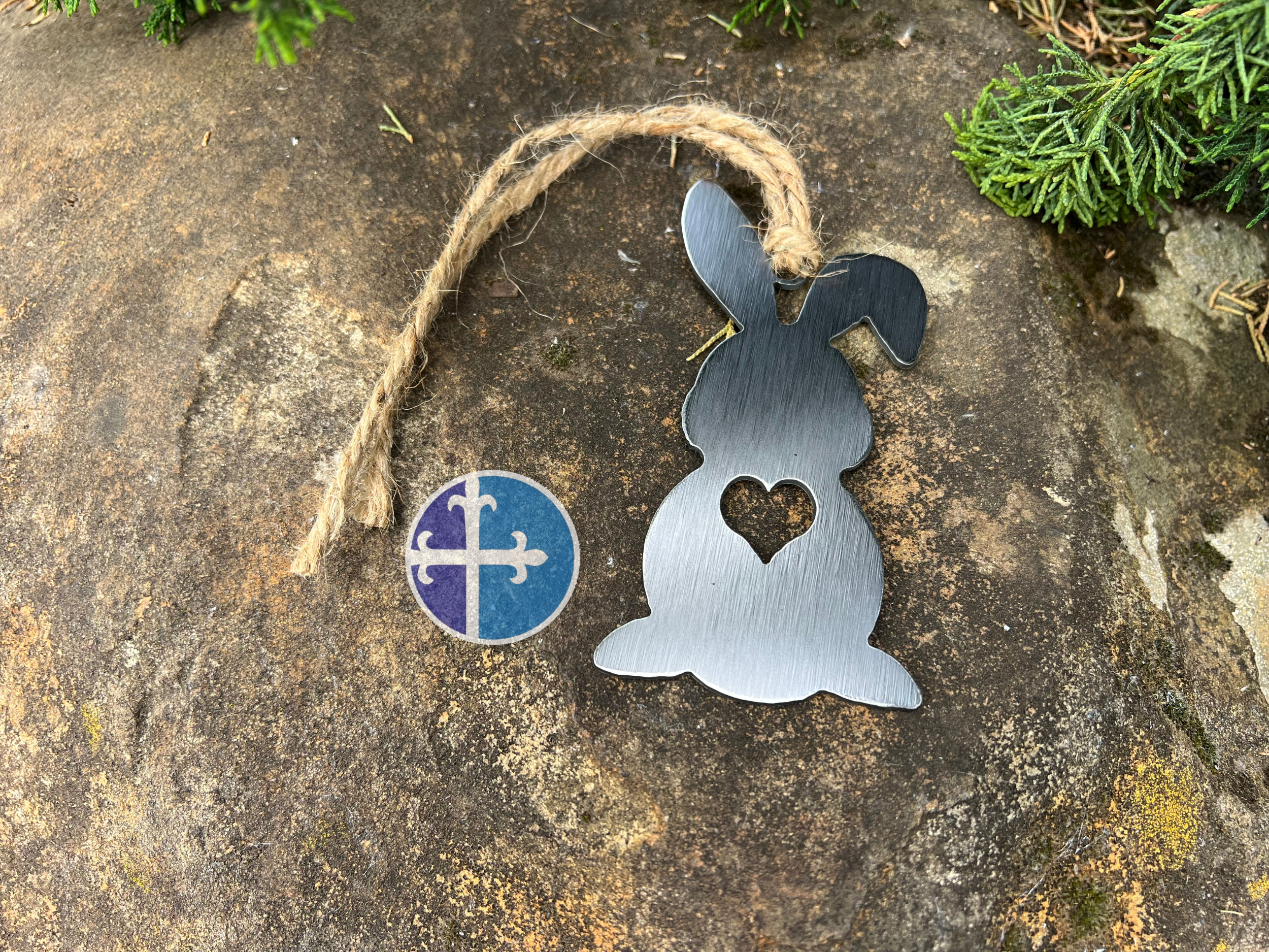 Steel bunny silhouette ornament with a heart cutout, hanging from jute twine, displayed on a stone background with greenery.