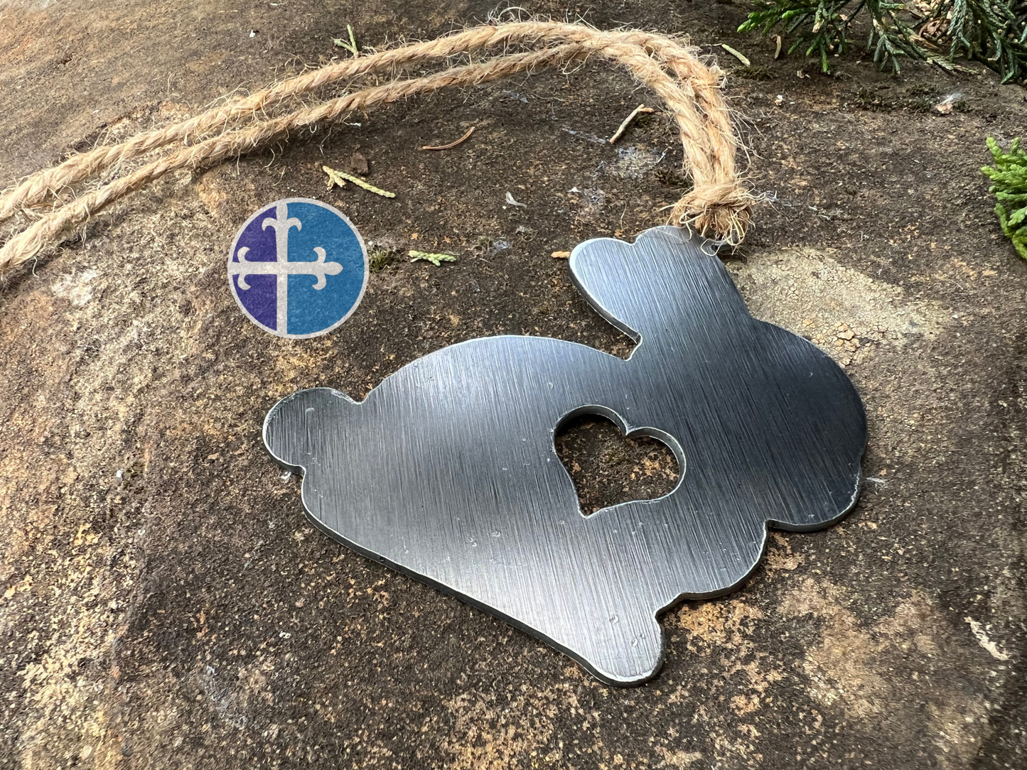 A metal bunny ornament with a heart cutout hanging on a rustic jute string, displayed on a stone background.
