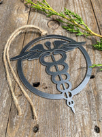 Load image into Gallery viewer, A steel ornament with a stylized design of a nursing emblem, featuring a serpent entwined rod, displayed on a rustic wooden background alongside a selection of natural stones and greenery.
