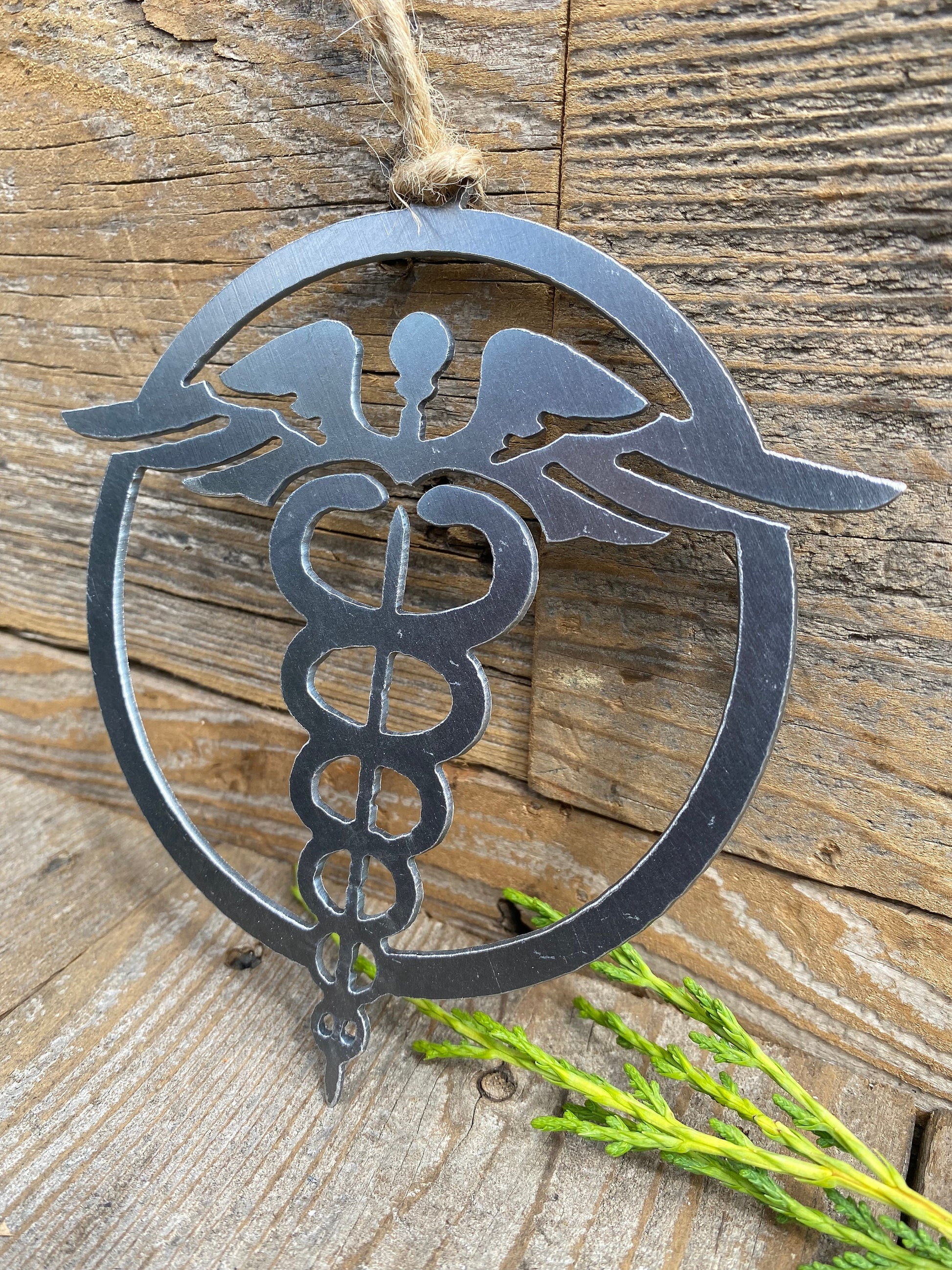 A steel ornament depicting a nurse's emblem, with a caduceus symbol in the center surrounded by an outer circle, hanging by a twine loop on a rustic wooden surface, with a sprig of greenery to the side.