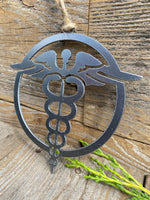 Load image into Gallery viewer, A steel ornament depicting a nurse&#39;s emblem, with a caduceus symbol in the center surrounded by an outer circle, hanging by a twine loop on a rustic wooden surface, with a sprig of greenery to the side.

