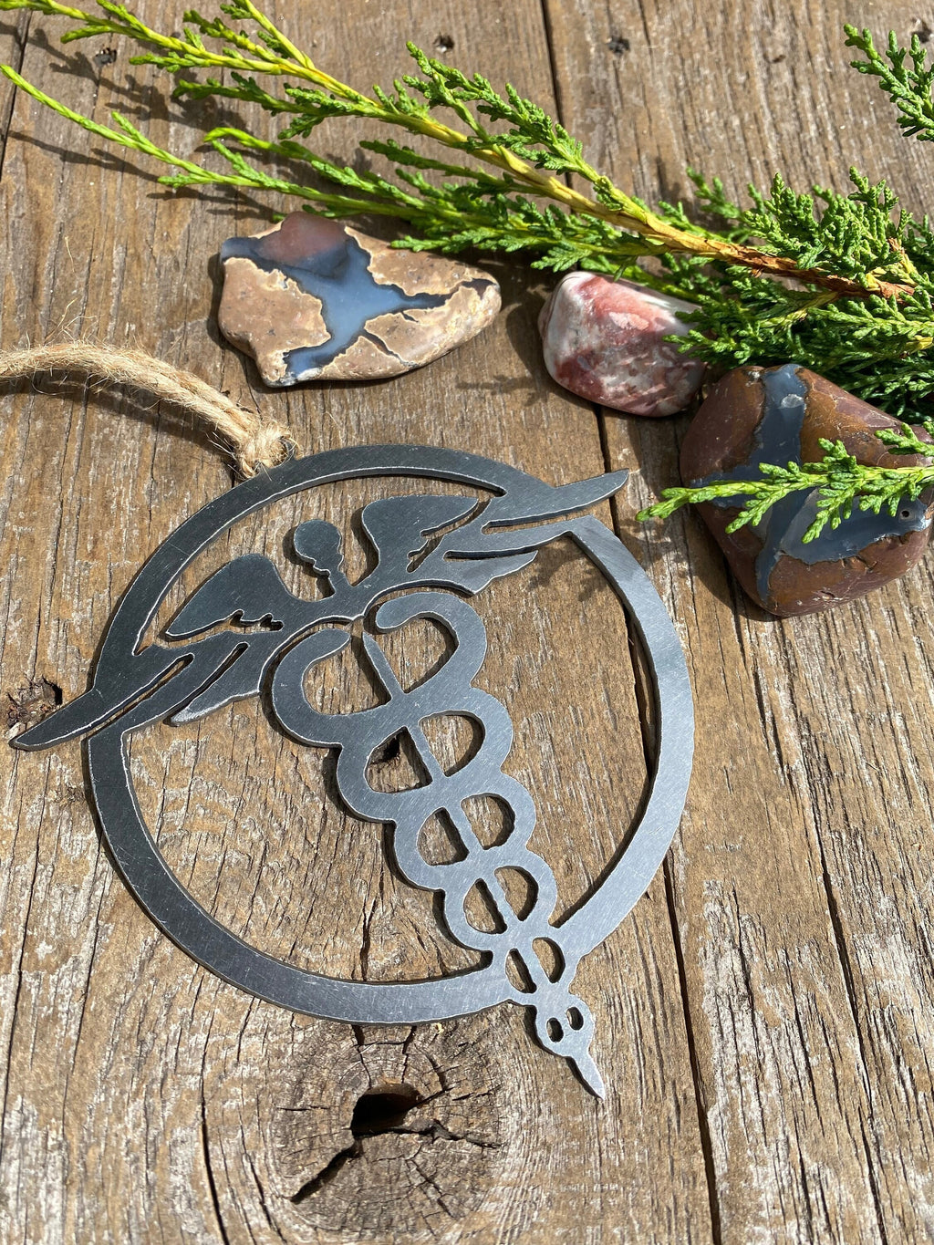 A steel ornament with a stylized design of a nursing emblem, featuring a serpent entwined rod, displayed on a rustic wooden background alongside a selection of natural stones and greenery.