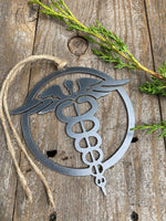 Load image into Gallery viewer, A steel ornament with a stylized design of a nursing emblem, featuring a serpent entwined rod, displayed on a rustic wooden background alongside a selection of natural stones and greenery.
