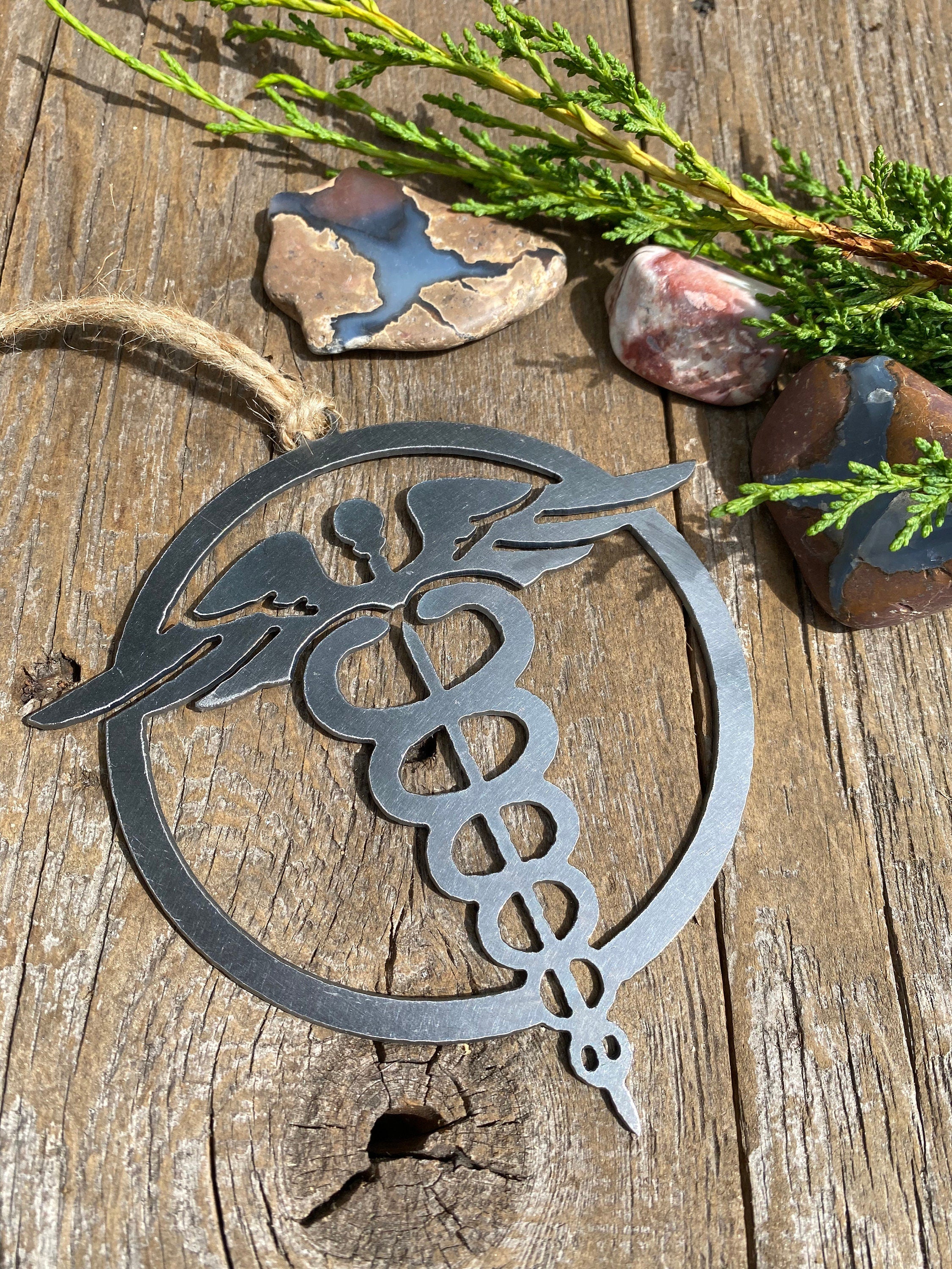 A steel ornament with a stylized design of a nursing emblem, featuring a serpent entwined rod, displayed on a rustic wooden background alongside a selection of natural stones and greenery.