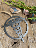 Load image into Gallery viewer, A steel ornament with a stylized design of a nursing emblem, featuring a serpent entwined rod, displayed on a rustic wooden background alongside a selection of natural stones and greenery.
